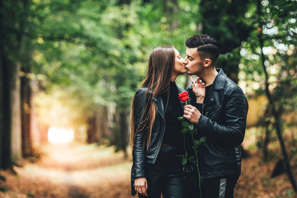 Passionate young man and woman kissing in the autumnal park, holding together a red rose. - Fotografie, Obrázek
