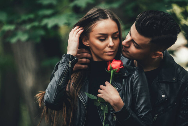 Brunette man embracing his girlfriend from behind with intention to kiss, while she holds a beautiful rose. - 写真・画像