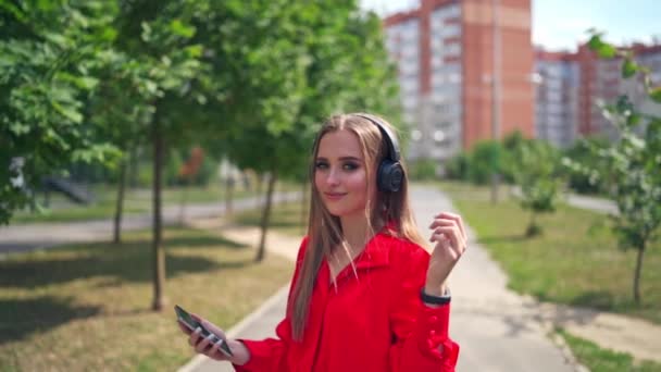 Lovely girl outdoors. Portrait of a young woman with headphones. Beautiful female listening to music in headphones on the city skyscrapers background. - Footage, Video