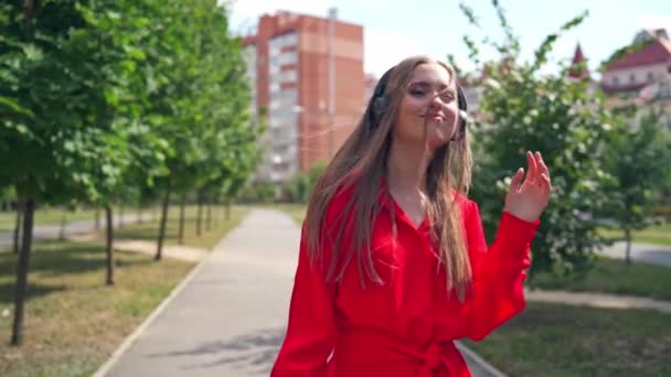 Beautiful young woman with headphones. Happy girl walking and enjoying listening to music in the street. Attractive female in red dress on the city background. - Кадри, відео