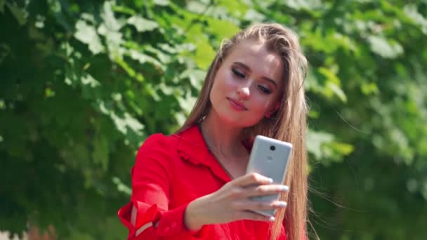 Attractive model with a mobile phone outdoors. Beautiful woman doing selfie in the street on green tree background. Lovely girl is recording video of herself on a phone. - Filmagem, Vídeo