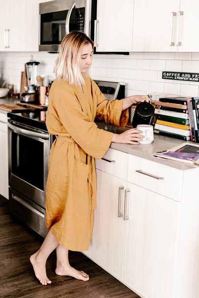Νεαρή γυναίκα φτιάχνει τον πρωινό της καφέ. Πρωινή ρουτίνα. Διαβάζω εφημερίδα. Λινό μπουρνούζι. Τρόπος ζωής στην κουζίνα. Λευκά ντουλάπια κουζίνας. Φωτεινή μοντέρνα κουζίνα. Millennial γυναίκα στο σπίτι της. Χαλαρώστε το χρόνο - Φωτογραφία, εικόνα
