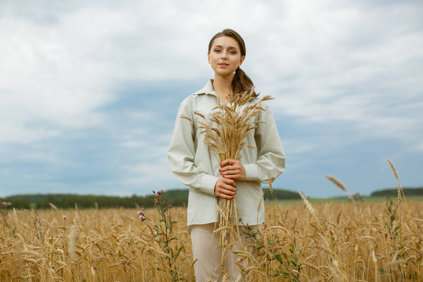 A lonely girl with an armful of ears stands in the middle of a wheat field. - Foto, Imagen