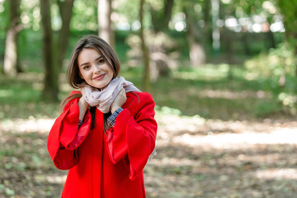 Image with adorable brunette girl touching her scarf, looking innocently and sincerely over blurred park background. - Photo, Image