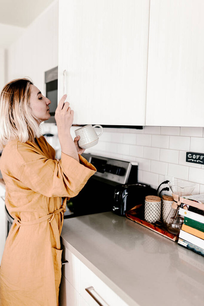 Молодая женщина готовит свой утренний кофе. Утренняя программа. Читаю газету. Постельный халат. Кухонный стиль жизни. Белые кухонные шкафы. Светлая современная кухня. Тысячелетняя женщина в своем доме.
 - Фото, изображение
