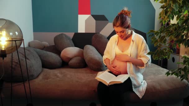 Νεαρή έγκυος γυναίκα χαϊδεύει την κοιλιά της και διαβάζει βιβλίο δυνατά. - Πλάνα, βίντεο