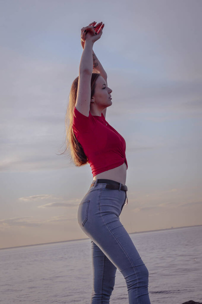 happy girl in a red blouse stretches on the shore by the sea. sunset, beautiful sea and sky - Foto, Imagem
