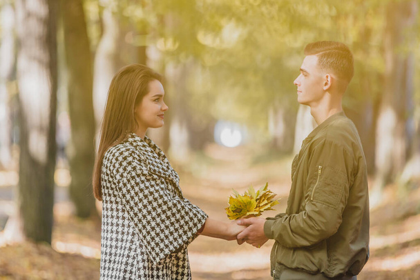 Real love. Beautiful young couple holding a bundle of leaves in hands and keeping eye contact on sunny autumn day in park. - Foto, afbeelding