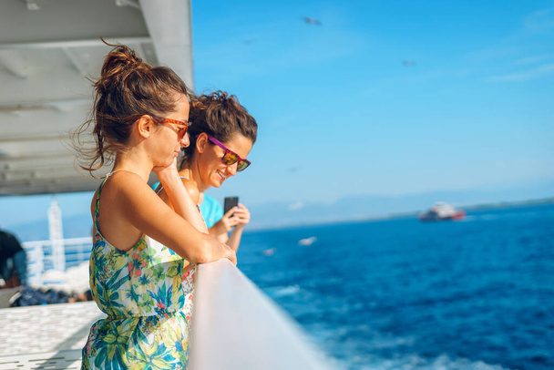 Dos jóvenes amigas hermanas de pie junto a la valla en la cubierta del ferry o barco navegando hacia el destino turístico de la isla en vacaciones de verano mirando al mar en un día soleado
 - Foto, imagen