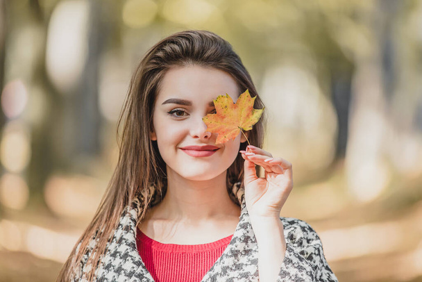 Close-up photo of lovely caucasian girl hiding one eye behind red-yellow autumn maple leaf, posing against blurred forest background. - Φωτογραφία, εικόνα
