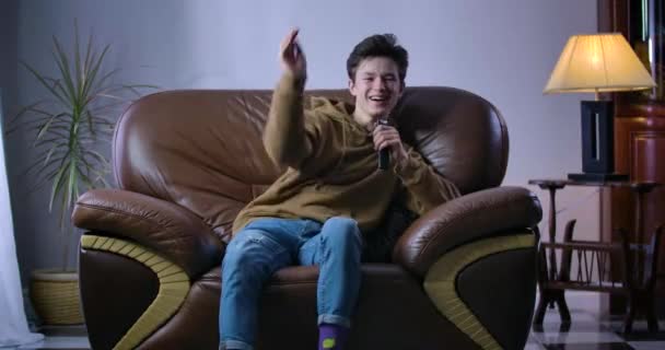Cheerful Caucasian boy laughing out loud as sitting on armchair with remote control. Joyful teenager watching comedy show or movie on TV at home. Leisure, relaxation, joy. Cinema 4k ProRes HQ. - Video, Çekim