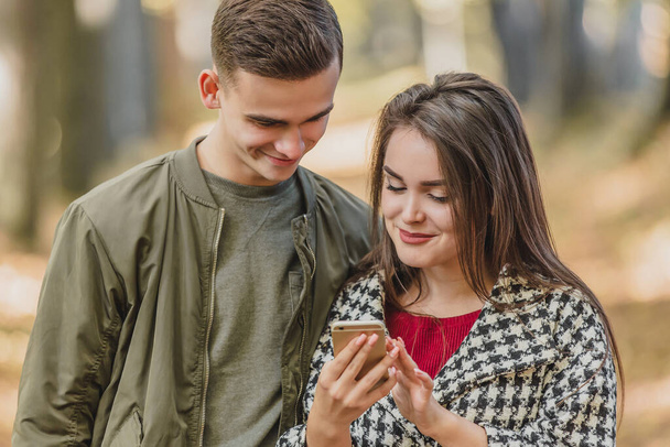 Close up photo of cheerful girl, showing photos to her boyfriend, while they are spending free time in autumn park. - Zdjęcie, obraz