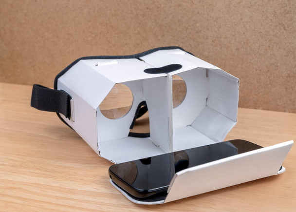A cardboard virtual reality headset. Front view, it is open a mobile phone on it. Lenses visible inside. It is resting on a wooden table next to a cork wall. - Photo, Image