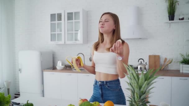 Slim woman with banana in the kitchen. Beautiful girl in the modern light kitchen. Young female eats sweet banana and throws banana peel on the floor. - Filmati, video