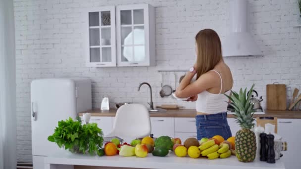 Vista lateral de una mujer bonita en la cocina. Mesa de cocina llena de frutas y verduras orgánicas frescas para una dieta saludable. Mujer comiendo comida dietética en casa
. - Metraje, vídeo
