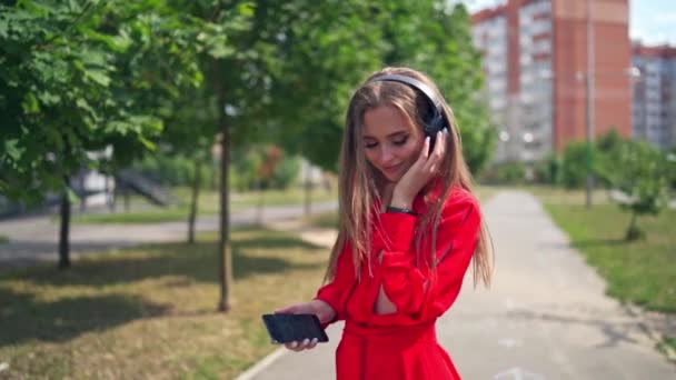 Attractive woman listening to music outdoors. Young female in red dress with headphones enjoys music in the city street in summer. - Metraje, vídeo