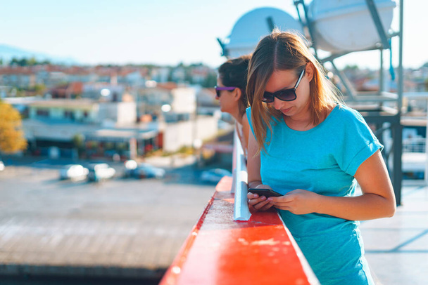 Νεαρή γυναίκα που χρησιμοποιεί κινητό τηλέφωνο ενώ βρίσκεται στο κατάστρωμα του πλοίου ή του πλοίου στέλνοντας γραπτό μήνυμα ή διαβάζοντας χρησιμοποιώντας εφαρμογή ή GPS πλοήγησης - Φωτογραφία, εικόνα