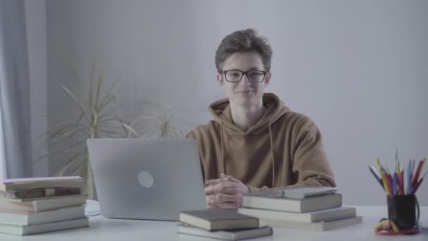 Confident genius boy in eyeglasses posing indoors. Portrait of intelligent Caucasian teenager sitting at the table with laptop and books and smiling at camera. Lifestyle, confidence, intelligence. - Imágenes, Vídeo
