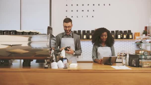 Hipster caucasian barista with beard and african young woman working in modern trendy coffee shop cafe, man washing cup and woman using laptop. Concept Preparing in for opening Small Business and - Video, Çekim