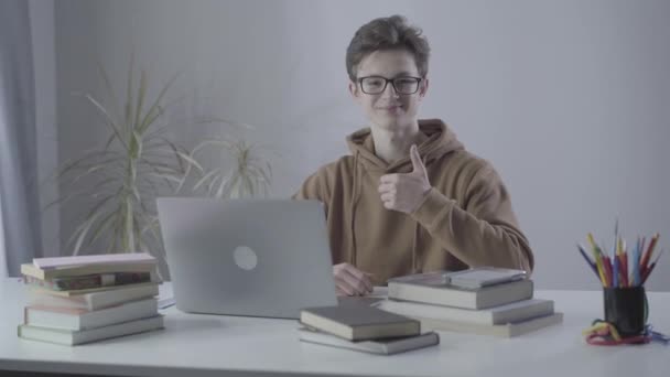 Positive college student showing thumb up and smiling at camera. Portrait of smart Caucasian brunette boy in eyeglasses sitting at the table with laptop. Intelligence, education, diligence. - Кадри, відео