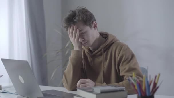 Tired millennial college student rubbing face as sitting at the table with laptop. Portrait of tired exhausted Caucasian brunette boy doing homework. Education, intelligence, adolescence, lifestyle. - Filmagem, Vídeo