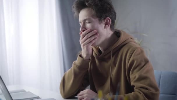 Side view close-up of tired millennial boy yawning and stretching. Portrait of exhausted Caucasian young brunette boy doing homework sitting at the table with laptop and books. Education concept. - Materiał filmowy, wideo
