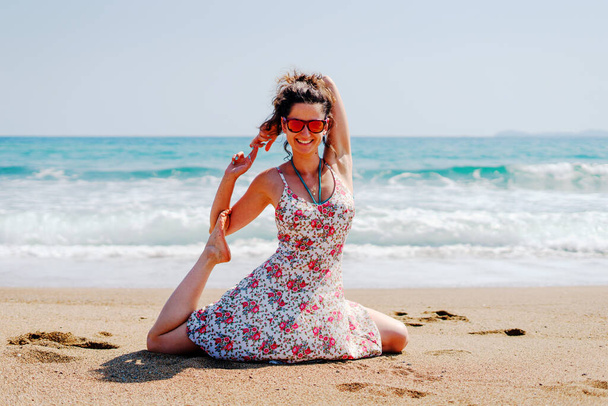 Fiatal nő nyári ruha gyakorlat jóga a strandon a tenger vagy az óceán napsütéses nap nyújtózó galamb ázsiai helyzetben - Fotó, kép