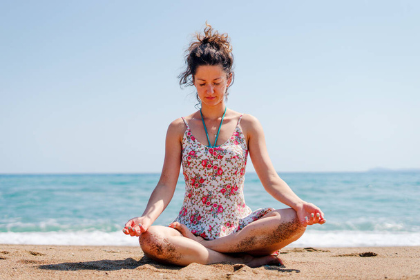 Молодая женщина в летнем платье практикует йогу медитируя на пляже у моря или океана в солнечный день растягивая медитацию расслабляя медитировать положение лотоса
 - Фото, изображение