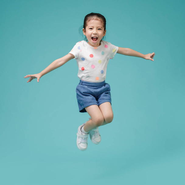 Speelse energieke Aziatische kleine meisje springen in de lucht, lege ruimte in studio schot geïsoleerd op kleurrijke blauwe achtergrond - Foto, afbeelding