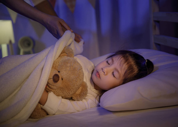 Madre mettendo coperta e accarezzando la testa per la sua bambina figlia sul letto in una camera da letto buia di notte, Bambino ragazza asiatica abbraccio orsacchiotto, Confortevole concetto di bambini a casa
 - Foto, immagini