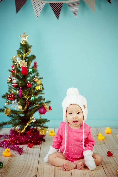 Little baby in knit winter clothing with knitted beanie and Decorating Christmas tree on green background - Foto, immagini