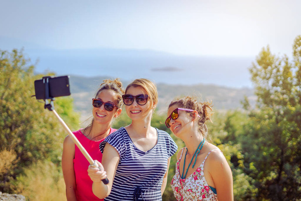 Yazlık elbiseli üç güzel bayan gezgin kız selfie çekerek akıllı telefonlarıyla selfie çekiyor arkadaş arkadaşlığı sonbaharda denizin yukarısındaki dağda. - Fotoğraf, Görsel