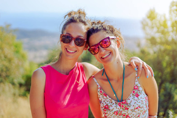 Two beautiful young woman female girl friends or sisters having fun in nature outdoors hugging embracing each other smiling wearing summer dress sunglasses in the autumn travel - 写真・画像