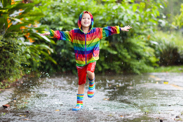 Gyerek játszik az esőben az őszi parkban. A gyerek sáros pocsolyába ugrik esős ősszel. Egy kisfiú esőkabátban és szivárványkabátban kint a szabadban, zuhanyozás közben. Gyerekek vízálló lábbeli és kabát. - Fotó, kép