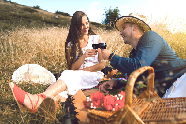 Young couple having a picnic on the blanket in the nature field on the mountain having a glasses of red wine celebrating love toasting on a date lovers happy in summer or autumn - Photo, Image