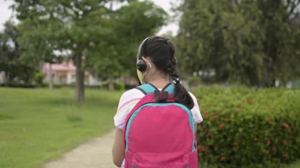 Zadní pohled na asijskou základní školačku v ochranné obličejové masce, která nosí sluchátka a chodí ve veřejném parku v obytné čtvrti. Žena teenager s batohem pomocí mobilního telefonu, zatímco ona je na cestě domů. Ochrana proti koronaviru. - Záběry, video