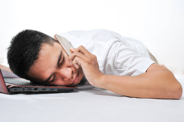 Sobretrabajado freelancer joven asiático durmiendo por ordenador portátil y teléfono en sus oídos en el trabajo cansado y sobrecargado, aislado sobre fondo blanco
 - Foto, Imagen