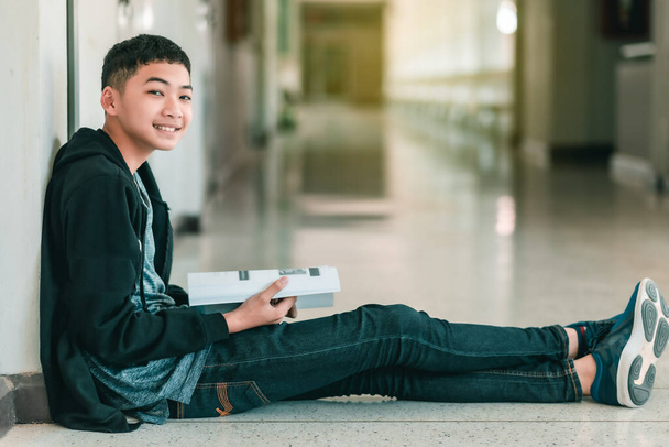 Un estudiante de secundaria masculino asiático está sentado en el pasillo de la escuela y usa una computadora portátil
. - Foto, imagen