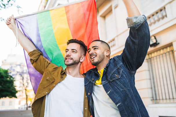 Portrait of young gay couple embracing and showing their love with rainbow flag at the street. LGBT and love concept - Fotó, kép