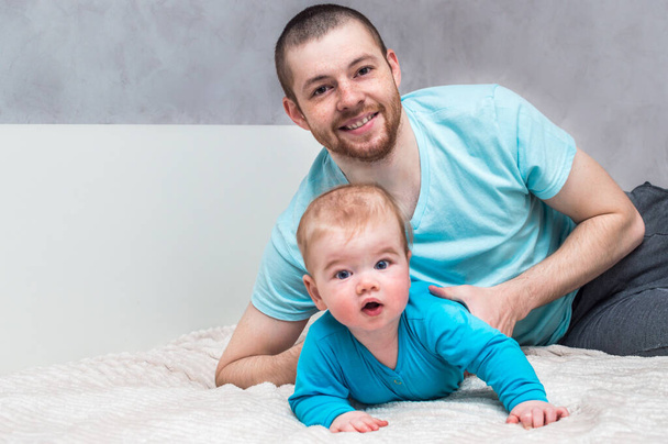 Portrait of a young smiling father with his infant in blue clothes on the bed in an embrace. Father's day concept. - 写真・画像