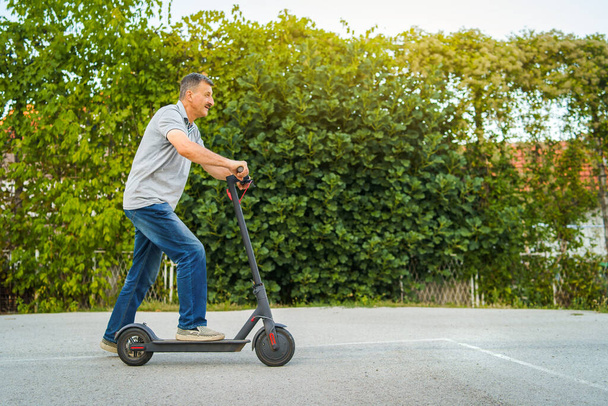 Hombre mayor montando patada empuje scooter en el asfalto delante de los arbustos cerca verde en un día de verano
 - Foto, Imagen