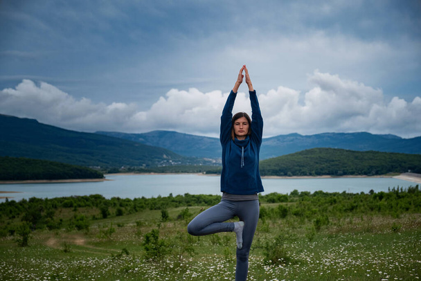 A young woman doing Hatha yoga in the field standing in yoga tree pose or asana Vrikshasana tre pose. Harmony with nature, Health and fitness concept. Self-analysis and soul-searching - Photo, Image