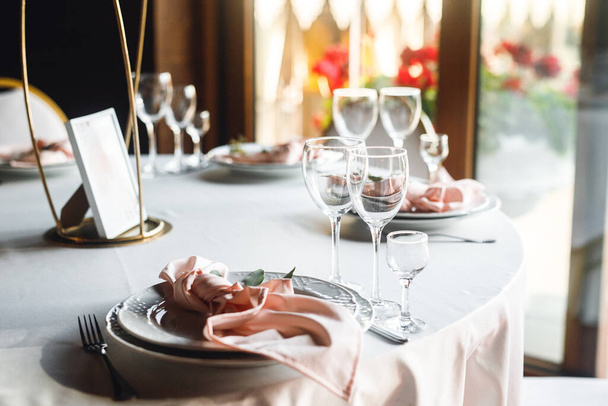 Ünnepi asztalok bezárása üres borospoharakkal és tányérral. Ünnepi bankett fehér tányérokkal, rózsaszín szalvétával és borosüveggel fehér és vörösborhoz a fehér asztalon az étteremben - Fotó, kép