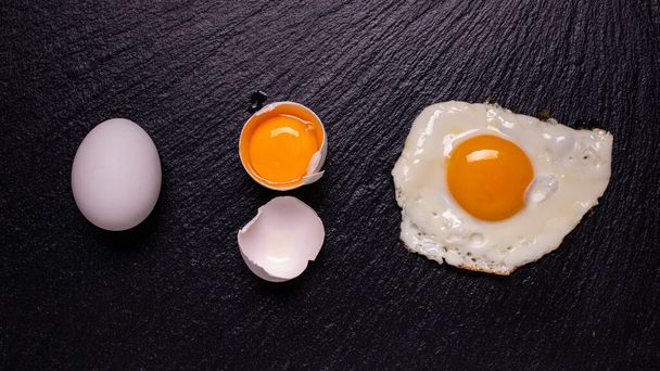 on the black slate stone, with a top view, in sequence, a white egg, an egg yolk in the shell and a fried egg. Still life - Foto, Imagen