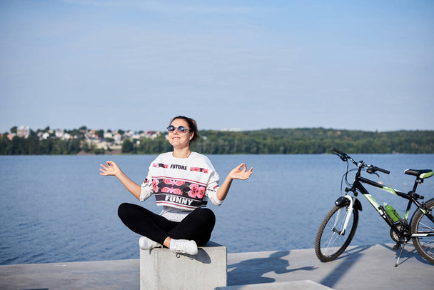 Sep 7, 2019-Ternopil/Ukraine:Young brunette woman, wearing black leggings and white sweatshot, sitting, resting on concrete platform by city lake with bicycle aside. Sports bike ride in summer morning - 写真・画像