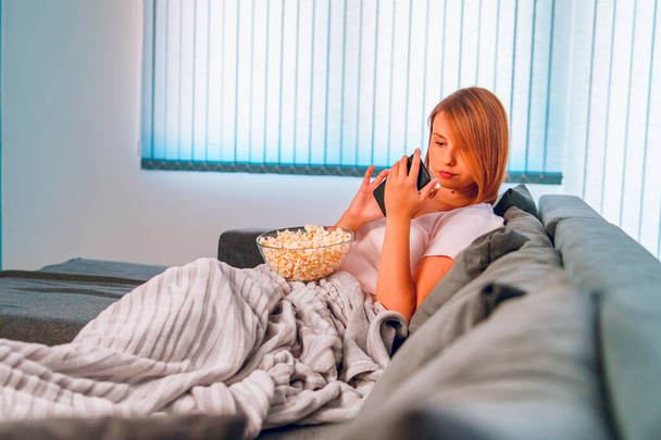 Young woman sitting on the bed sofa at home, lying with a popcorn bowl covered with blanket at her apartment alone enjoying using mobile smart phone to take selfies photo or to send sms message - Photo, Image