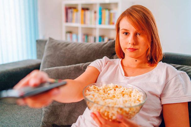 Young woman sitting on the bed sofa at home, lying with a popcorn bowl watching TV covered with blanket at her apartment alone enjoying movies or series holding remote control pointed - Photo, Image