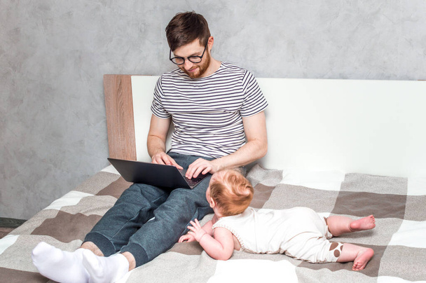 Young man working on a laptop in bed at home and baby beside. Concept Working remotely - Photo, image