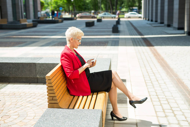 beautiful stylish woman in a red jacket sits on a bench with a tablet - Фото, изображение
