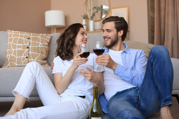 Happy couple sitting, relaxing on floor in living room, drinking red wine. Smiling young husband and wife rest at home enjoy romantic date on family weekend together. - Фото, изображение